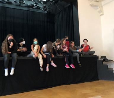 Mehrere Mädchen* sitzen auf einer Bühne. Eine von ihnen spricht ins Mikrophon.